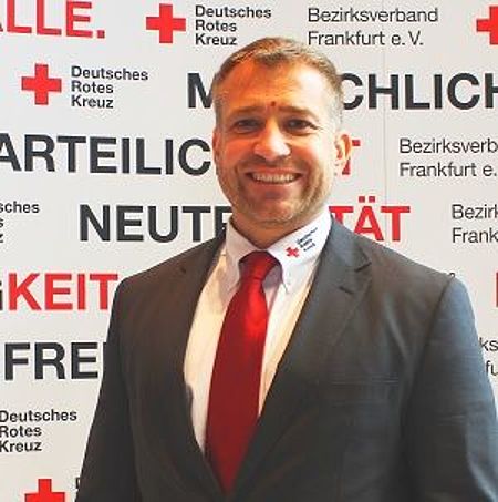 Marco Schmitz DRK Frankfurt am Main e.V. Das Bild zeigt eine Nahaufnahme von Marco Schmitz vor einer Wand mit Logo und Claims des DRK Frankfurt am Main